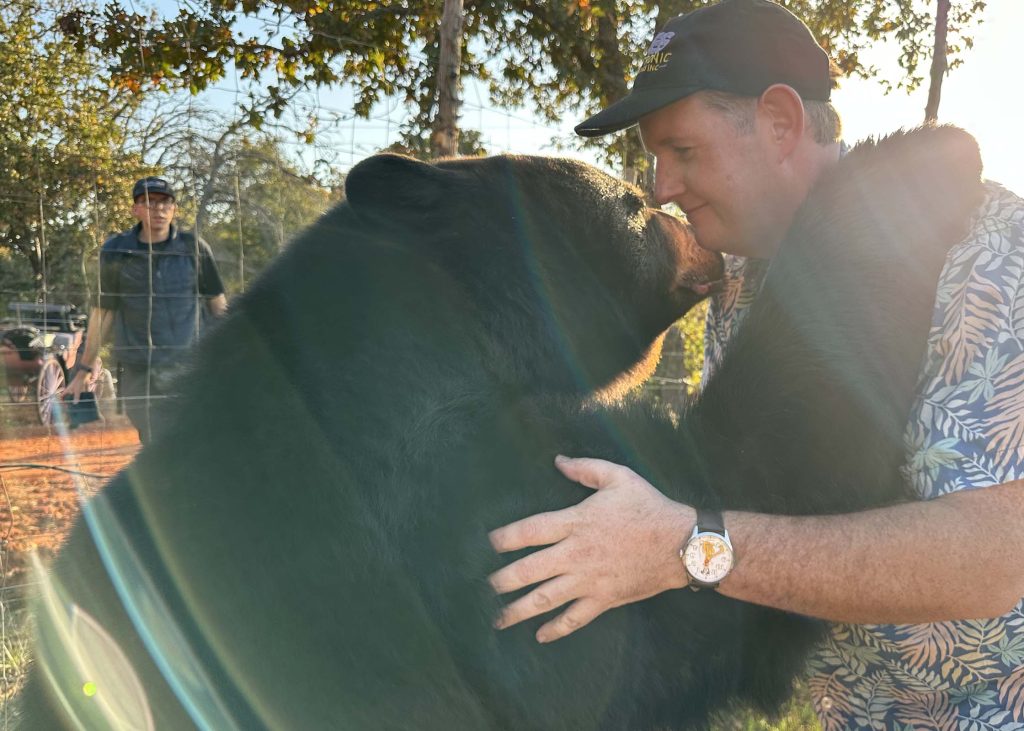 Hollywood Bear shares an outdoor greeting with a man beside the black bear character as crew stand nearby.